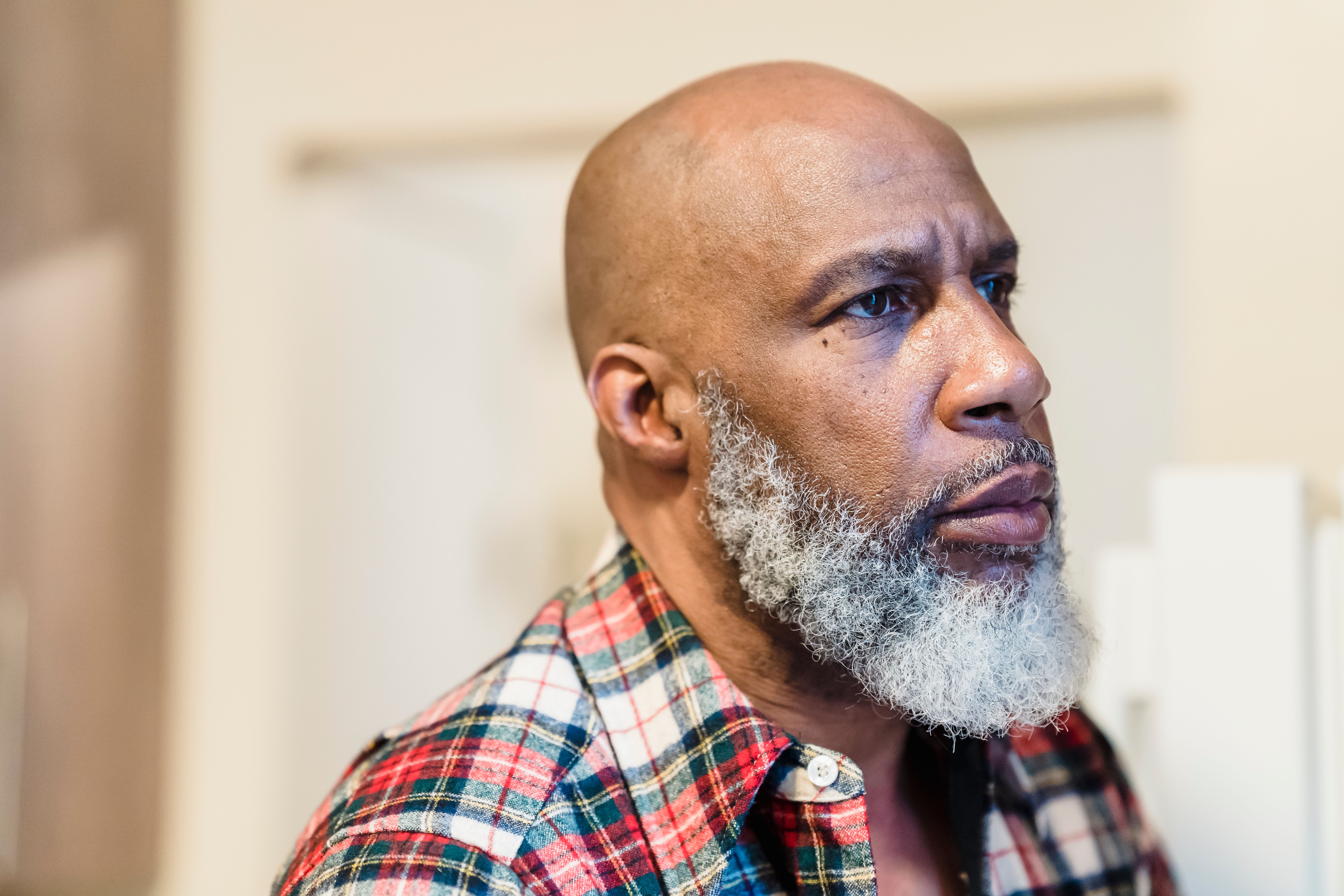 A thoughtful portrait of a bald, bearded man in a plaid shirt, looking away indoors.